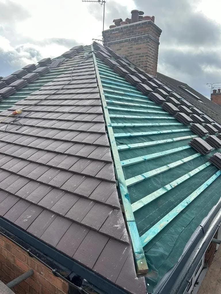 The hip roof half stripped: old tiles still along one side, fresh breathable membrane and battens on the other, meeting at the chimney stack with its pots.