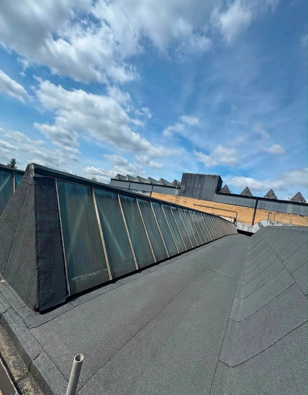 The finished factory roof: fresh grey mineral-surfaced cap sheet across the slopes, with new black detailing wrapped around the long run of glazed roof lanterns and the saw-tooth factory roofline behind.