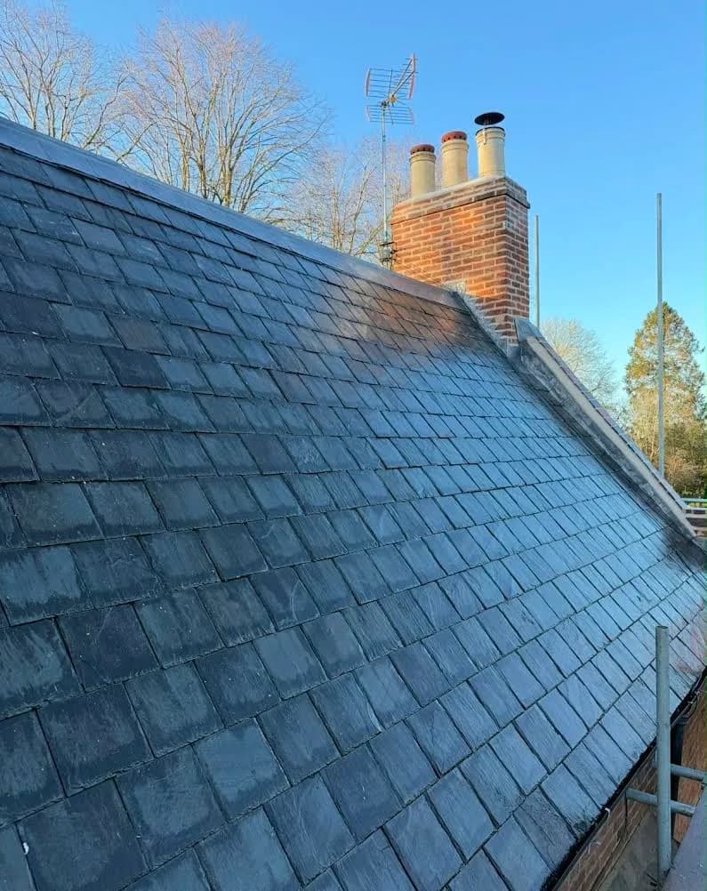 The finished roof in reclaimed Welsh slate, a long sweep of blue-grey slates running up to a lead ridge along the apex, with a red-brick chimney stack carrying three cream pots and a television aerial against a clear winter sky.