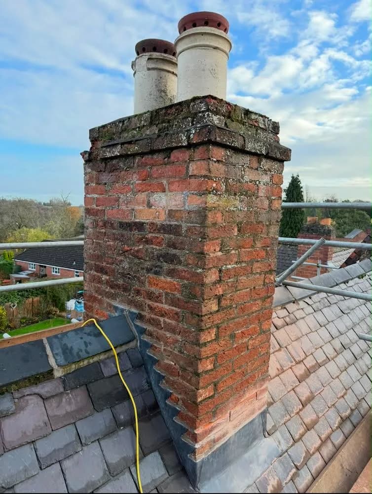 The chimney stack before repointing, its brickwork stained green with moss and algae and the mortar joints eroded back between the bricks, with two weathered cream pots on top and old slates around its base.