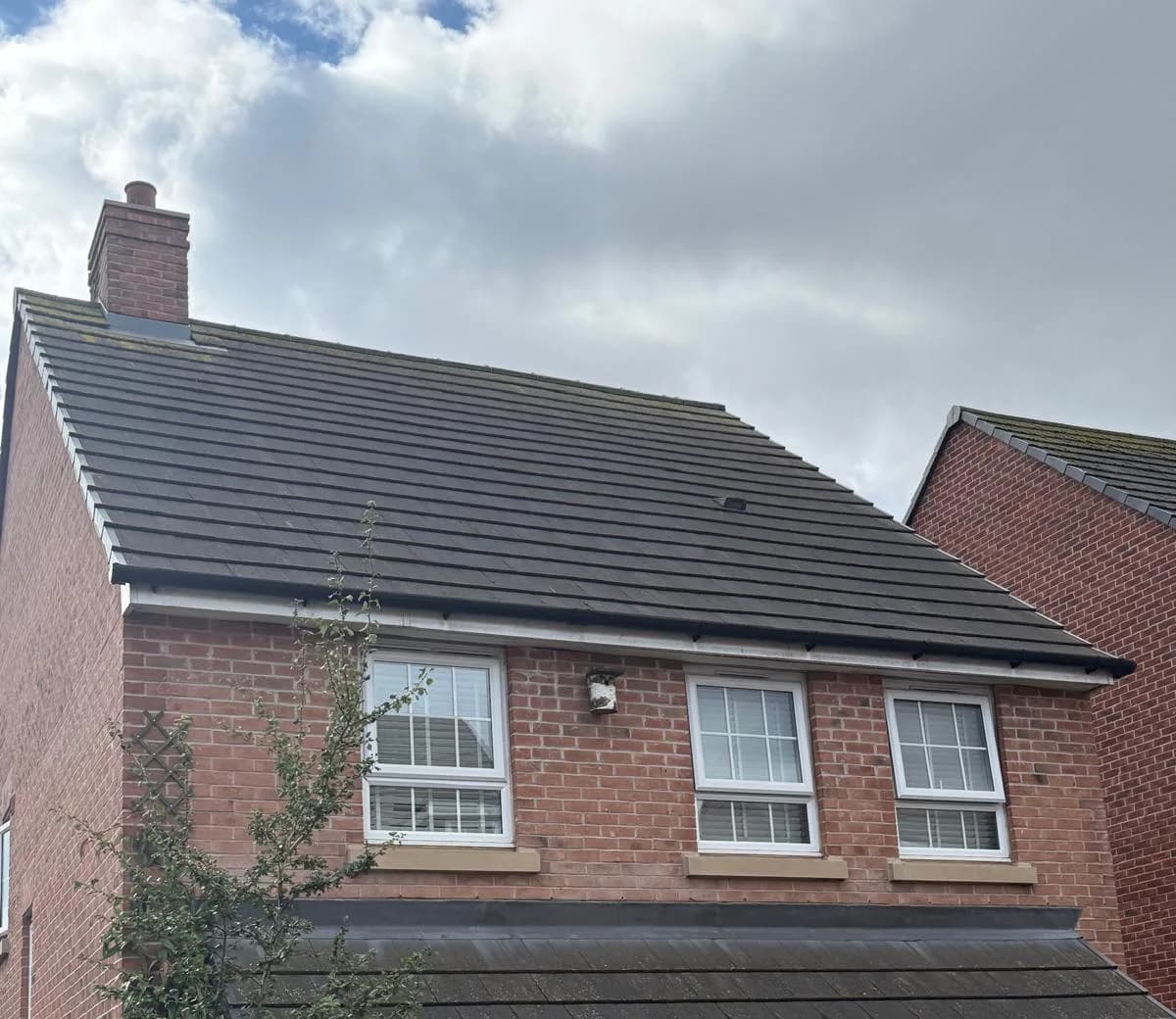 A red-brick detached house seen from the front at roof level, with a plain grey concrete-tiled roof, a brick chimney, upstairs windows and guttering along the eaves, and a neighbouring roof visible beyond.