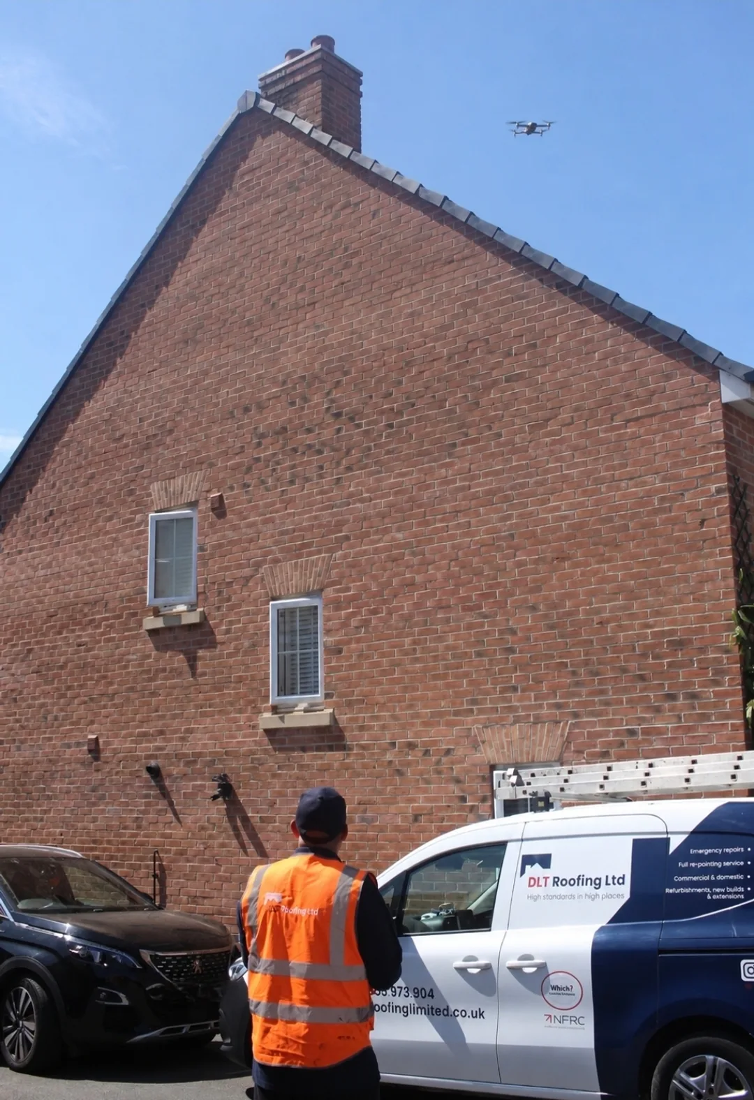 A small survey drone hovering in clear blue sky just above the ridge of a red-brick gable end, its chimney stack and dry verge visible, while a DLT Roofing surveyor in an orange hi-vis vest watches from the driveway beside the branded van.