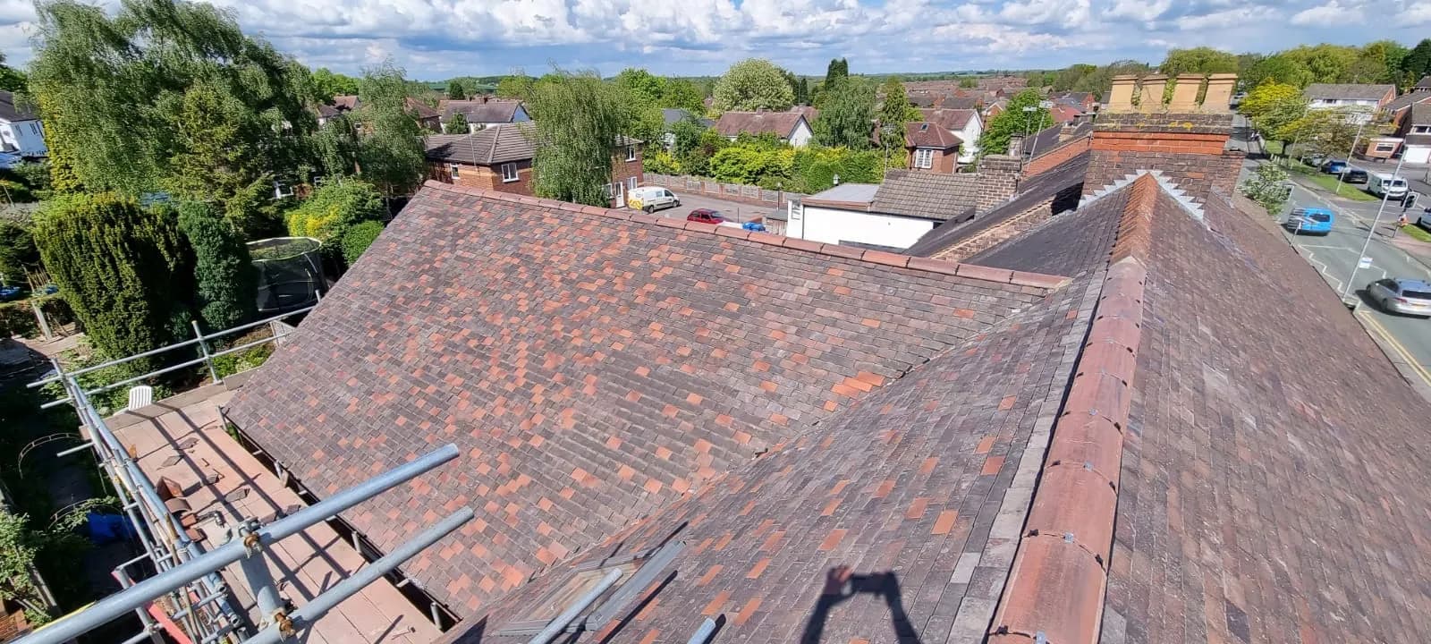 Aerial view across a large re-tiled roof of reddish-brown plain clay tiles, with suburban rooftops and greenery beyond.