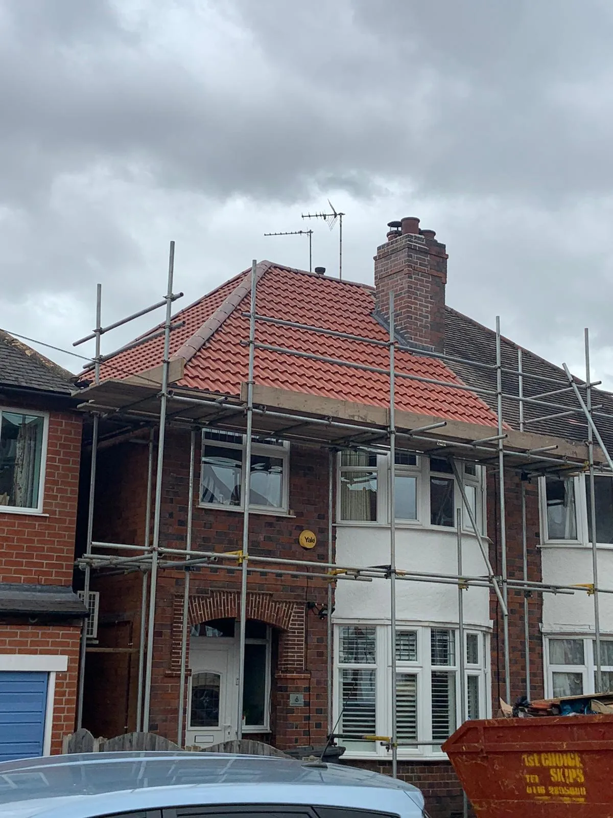 A semi-detached house mid re-roof, with a new red-tiled hip roof and chimney, surrounded by scaffolding and a skip in the driveway.